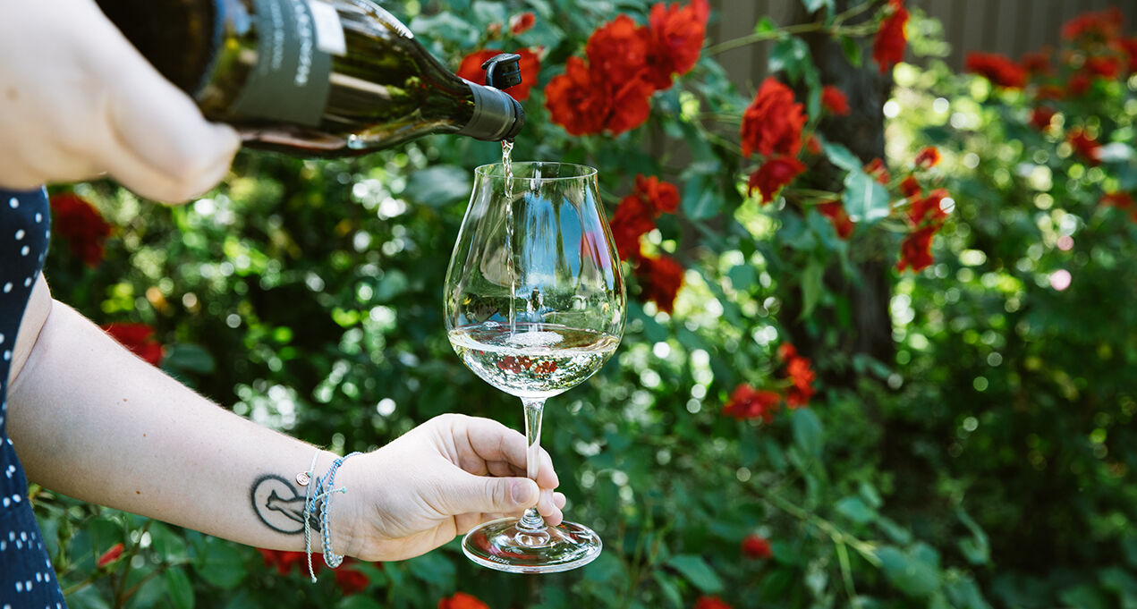 WillaKenzie Estate white wine being poured in a glass in front of red flowers.